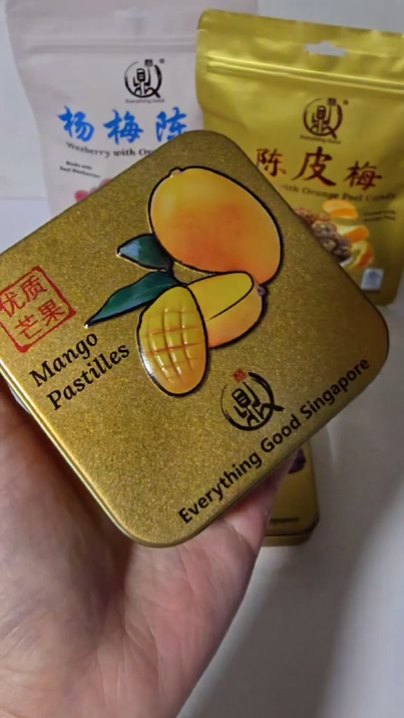 Hand holding an Everything Good Mango Mint Chewy Candy tin in front of Waxberry and Sour Plum candy pouches